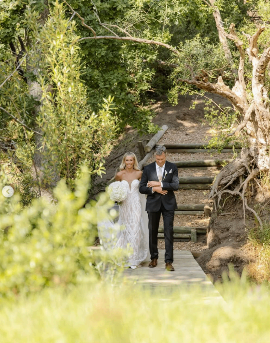 Bride walking with her father along a timber path framed by trees