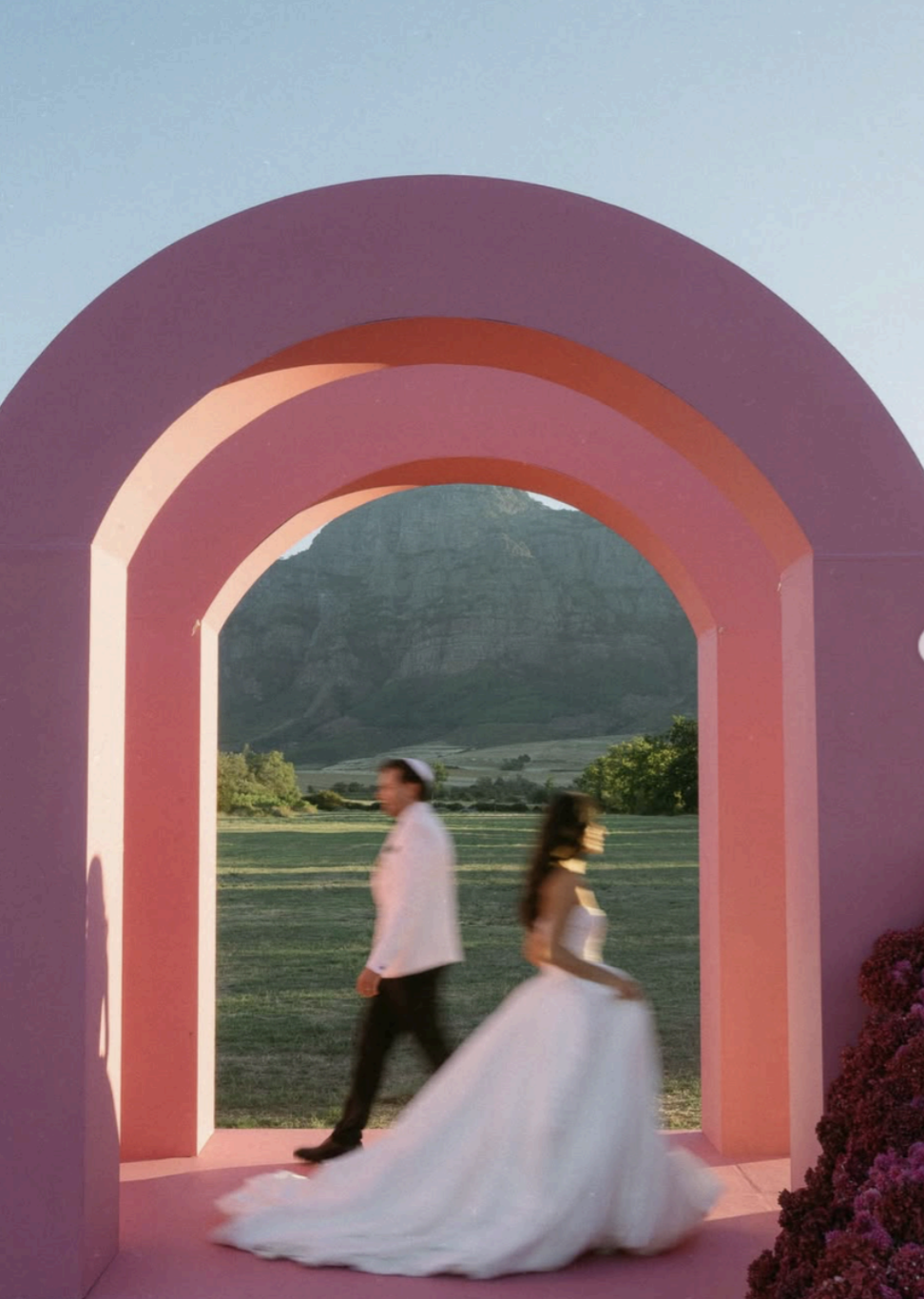 Bride and groom walking through a sculptural pink archway with mountain backdrop